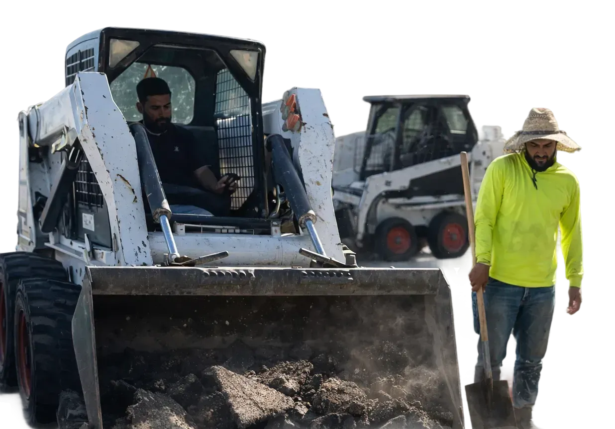 Construction worker in a straw hat and neon green shirt holding a shovel standing next to a skid steer loader filled with dirt and operated by a seated man.
