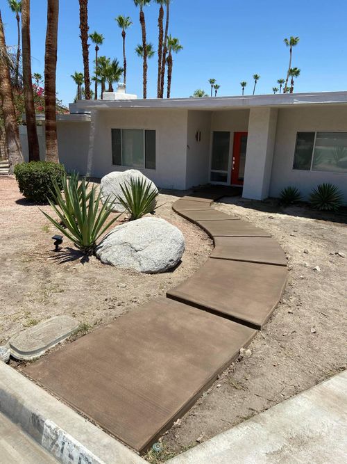 Curved concrete walkway leading to the front entrance of a single-story home with desert landscaping and palm trees.