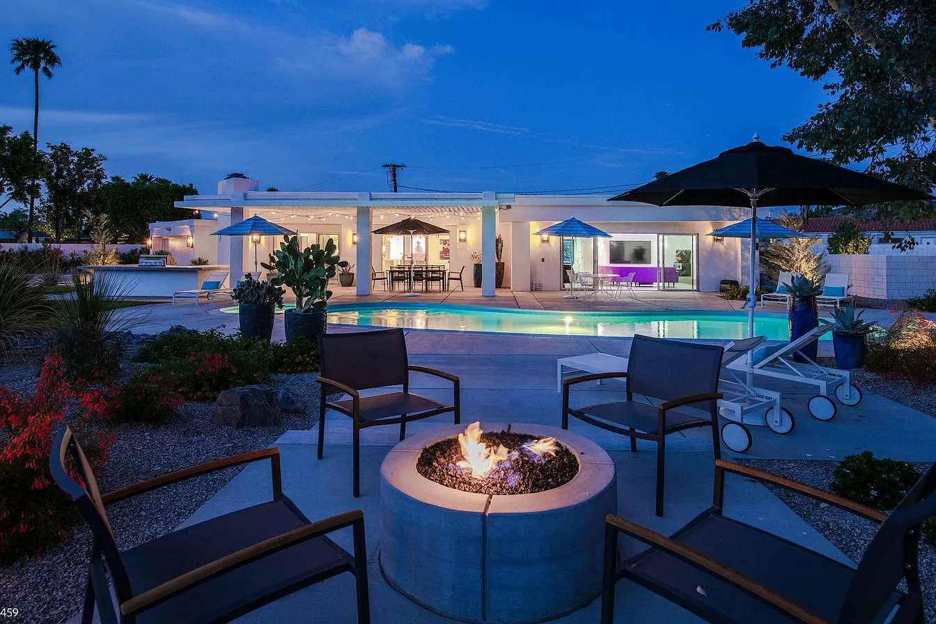 Outdoor patio with a modern fire pit surrounded by chairs, a swimming pool, and a contemporary house illuminated at dusk.