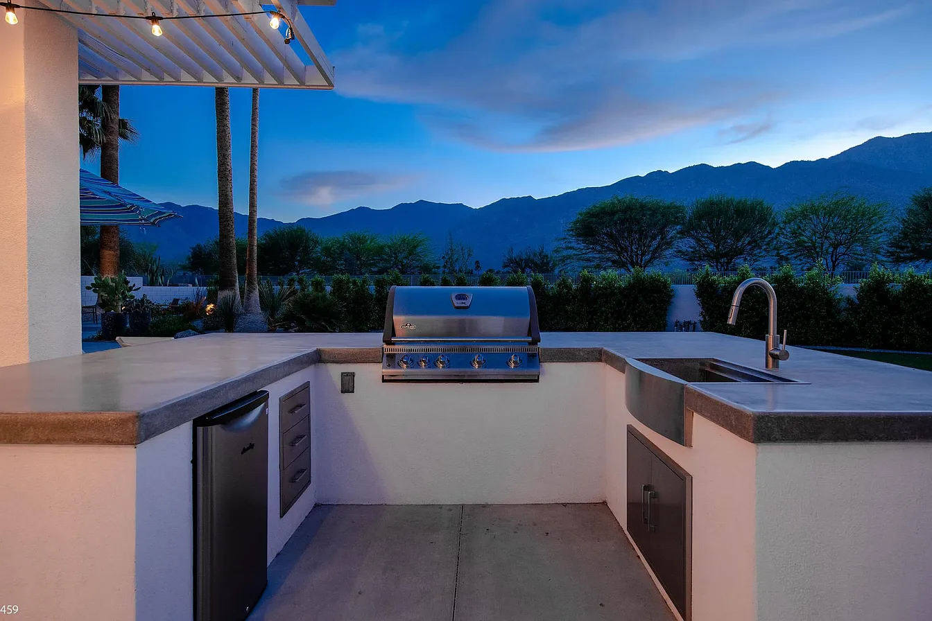 Outdoor kitchen with stainless steel grill, sink, mini fridge, and cabinets under an evening sky with mountains in the background.