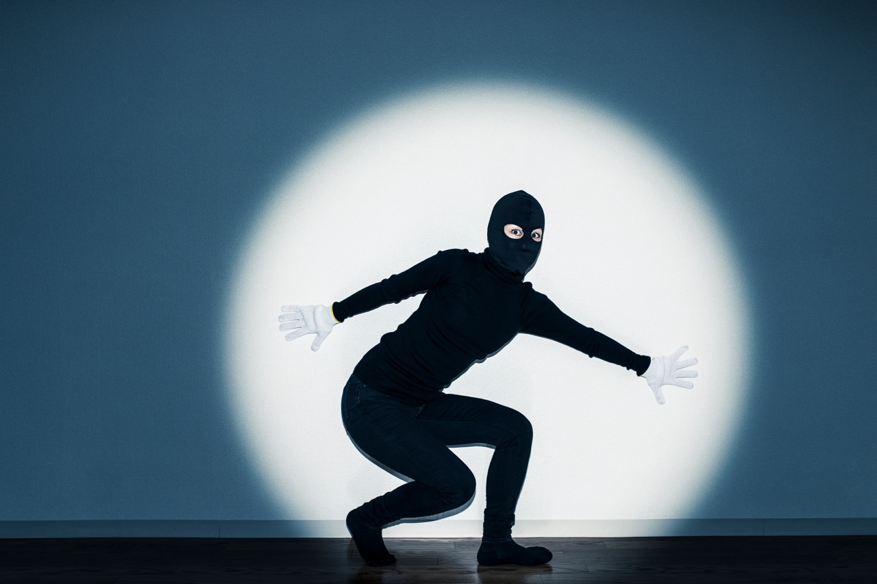 Person crouching in front of a wall spotlight, dressed in black with a balaclava and white gloves