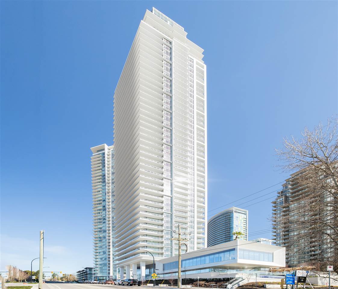 Tall modern residential building with balconies against a clear blue sky, surrounded by other high-rise buildings.