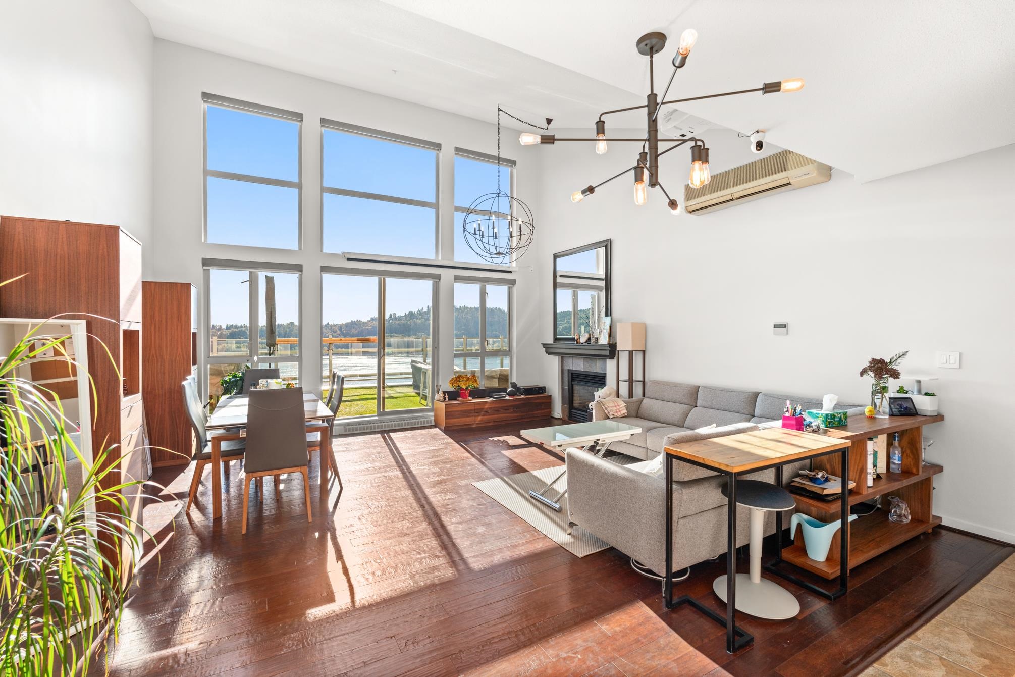 Bright living room with tall windows, wooden floor, dining table, gray sofa, modern chandeliers, and a fireplace.