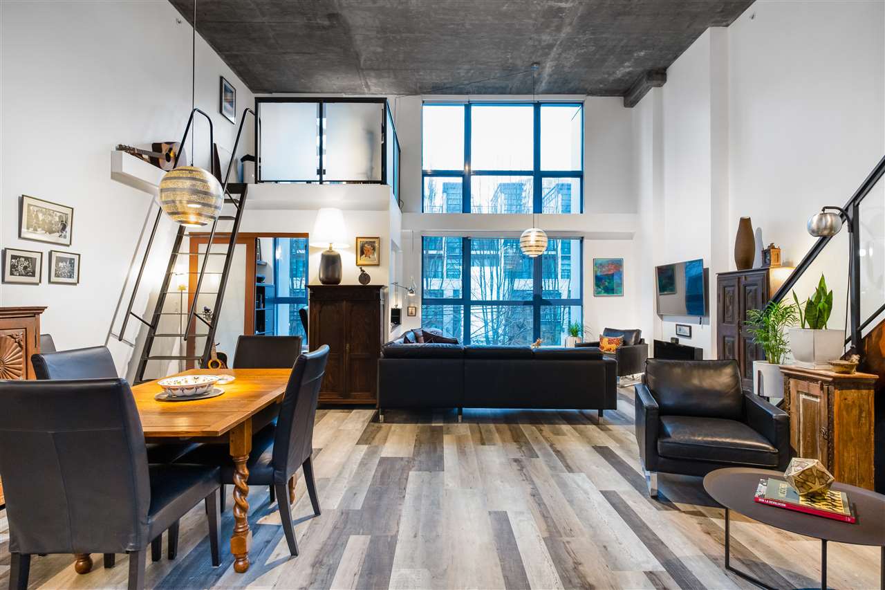 Modern loft-style living room with large windows, black leather seating, wooden dining table, and a mezzanine accessed by a ladder.
