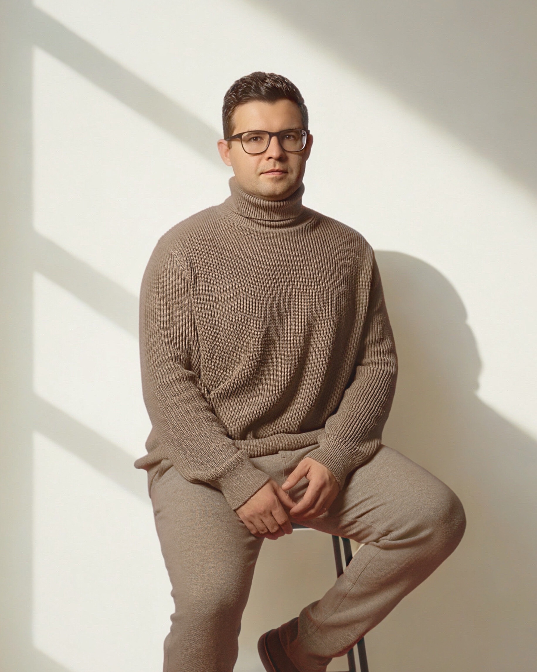Man wearing glasses and a brown turtleneck sweater sitting on a stool against a light background with shadows.