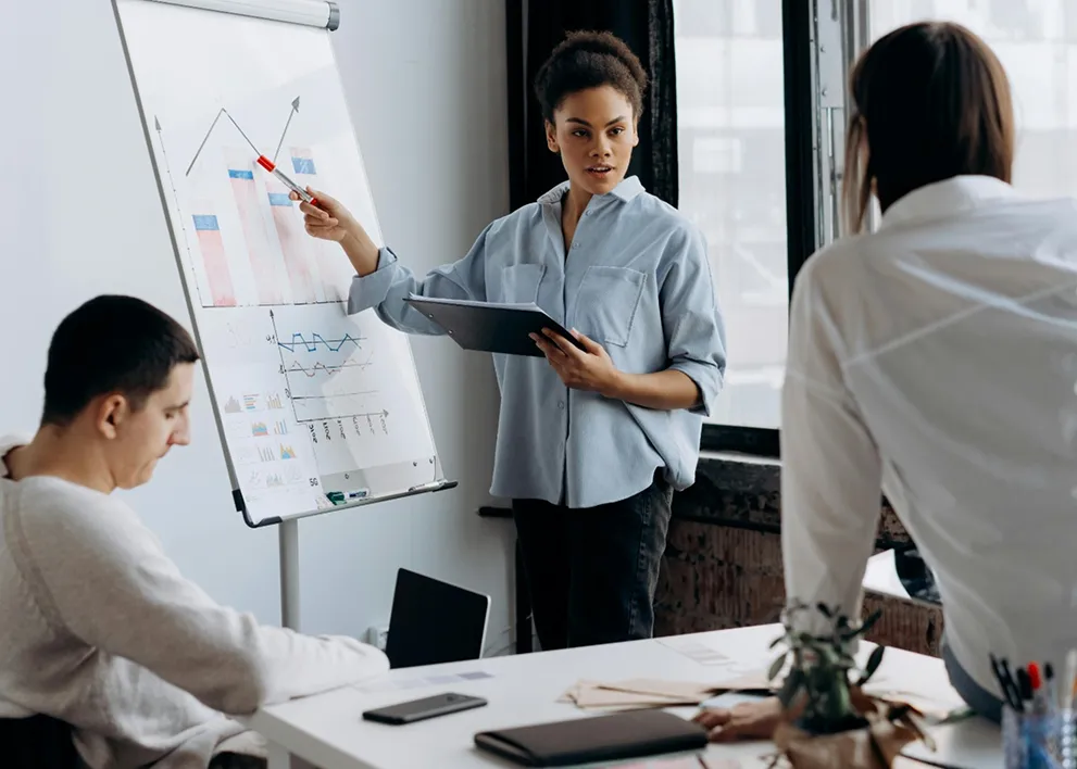 Woman presenting data charts on a flip chart to two seated colleagues in a modern office.