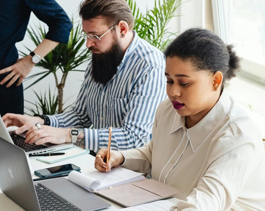 Two people focused on work; a woman writing notes beside an open laptop and a man typing on a laptop.