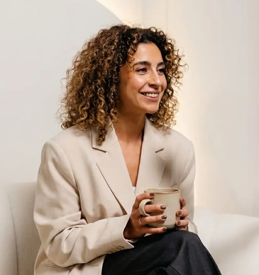 Smiling woman with curly hair wearing a beige blazer holding a ceramic mug while sitting on a white chair.