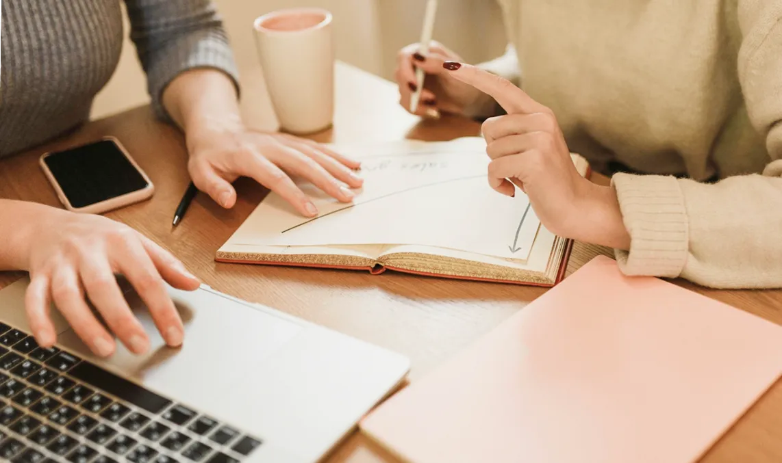 Two people working at a table with a laptop, an open notebook with a graph, a pen, and a cup.