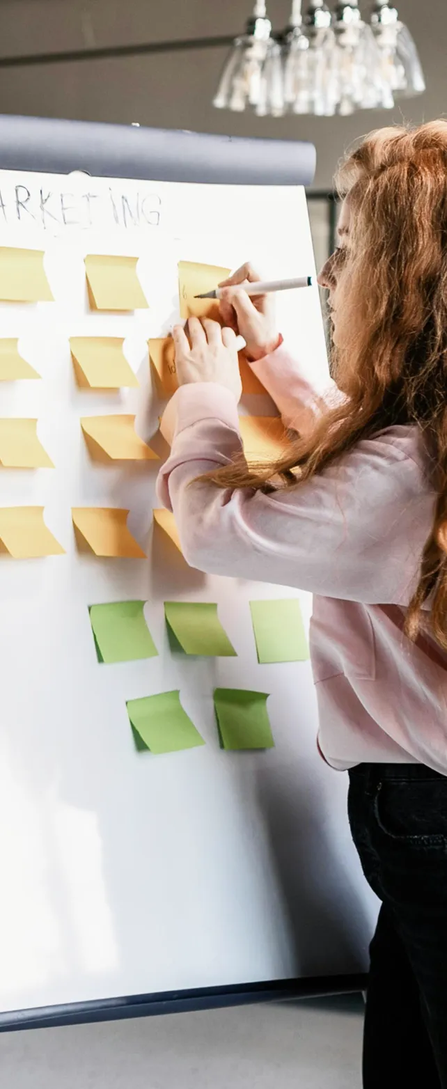 Woman with long curly hair writing on yellow sticky notes on a whiteboard titled marketing, with green sticky notes below.