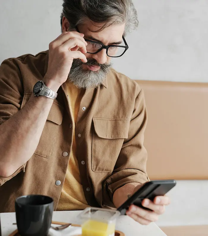Bearded man wearing glasses and a brown shirt adjusting his glasses while looking at a smartphone in a cafe setting.