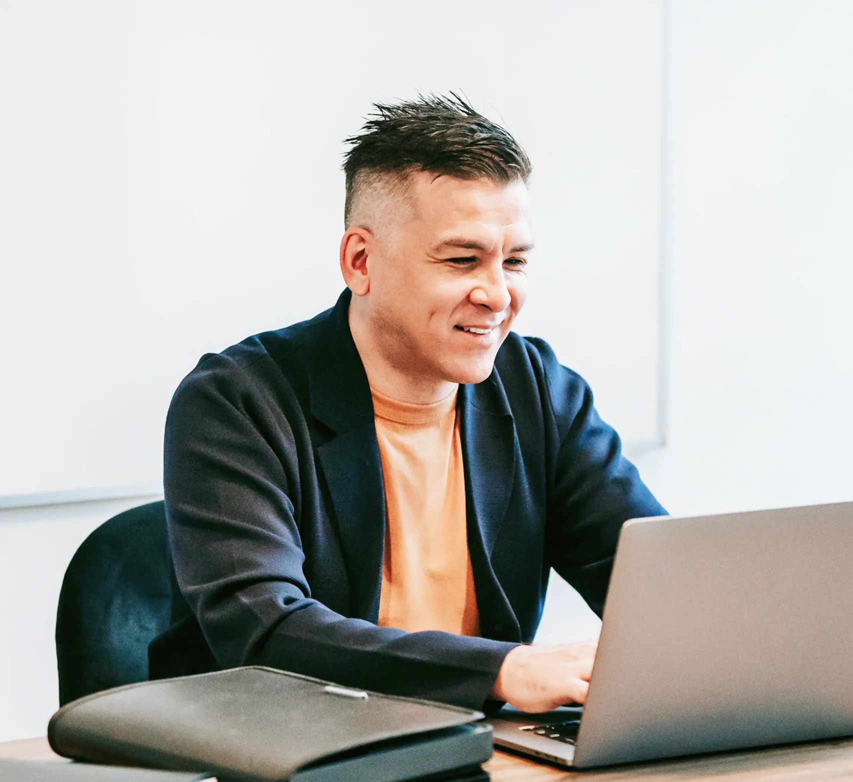 Man with short hair wearing a dark blazer and orange shirt, smiling while working on a laptop at a desk.