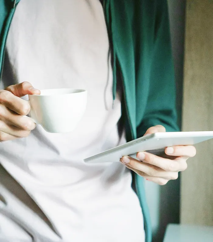 Person holding a white cup in one hand and a tablet in the other, wearing a light shirt and green jacket.