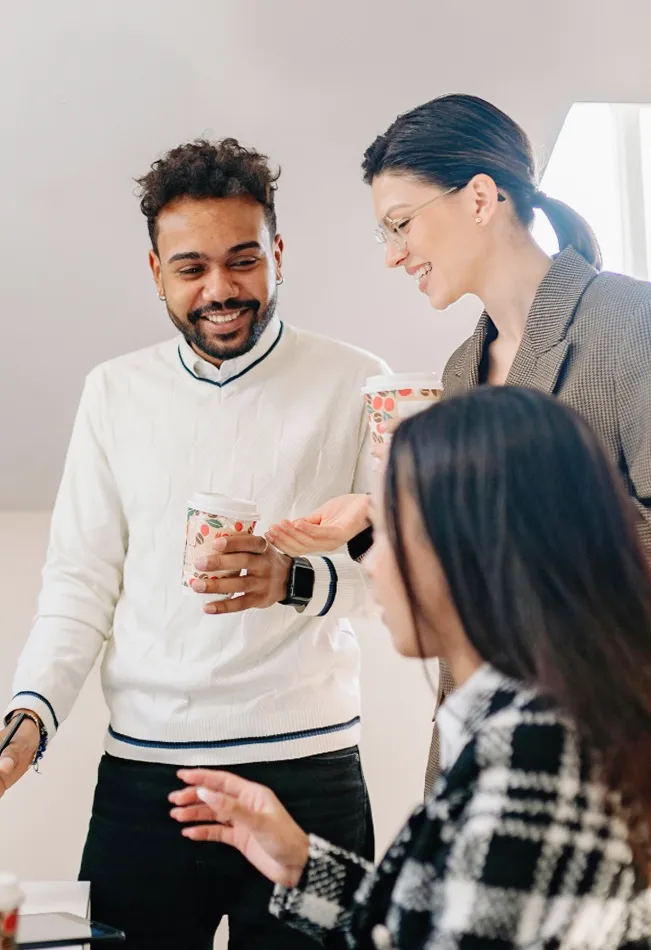 Two colleagues smiling and holding coffee cups while talking, with a third person blurred in the foreground.