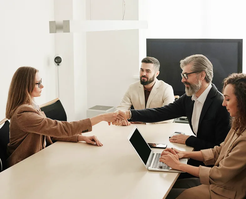 Two business professionals shaking hands across a conference table while two colleagues observe, one typing on a laptop.