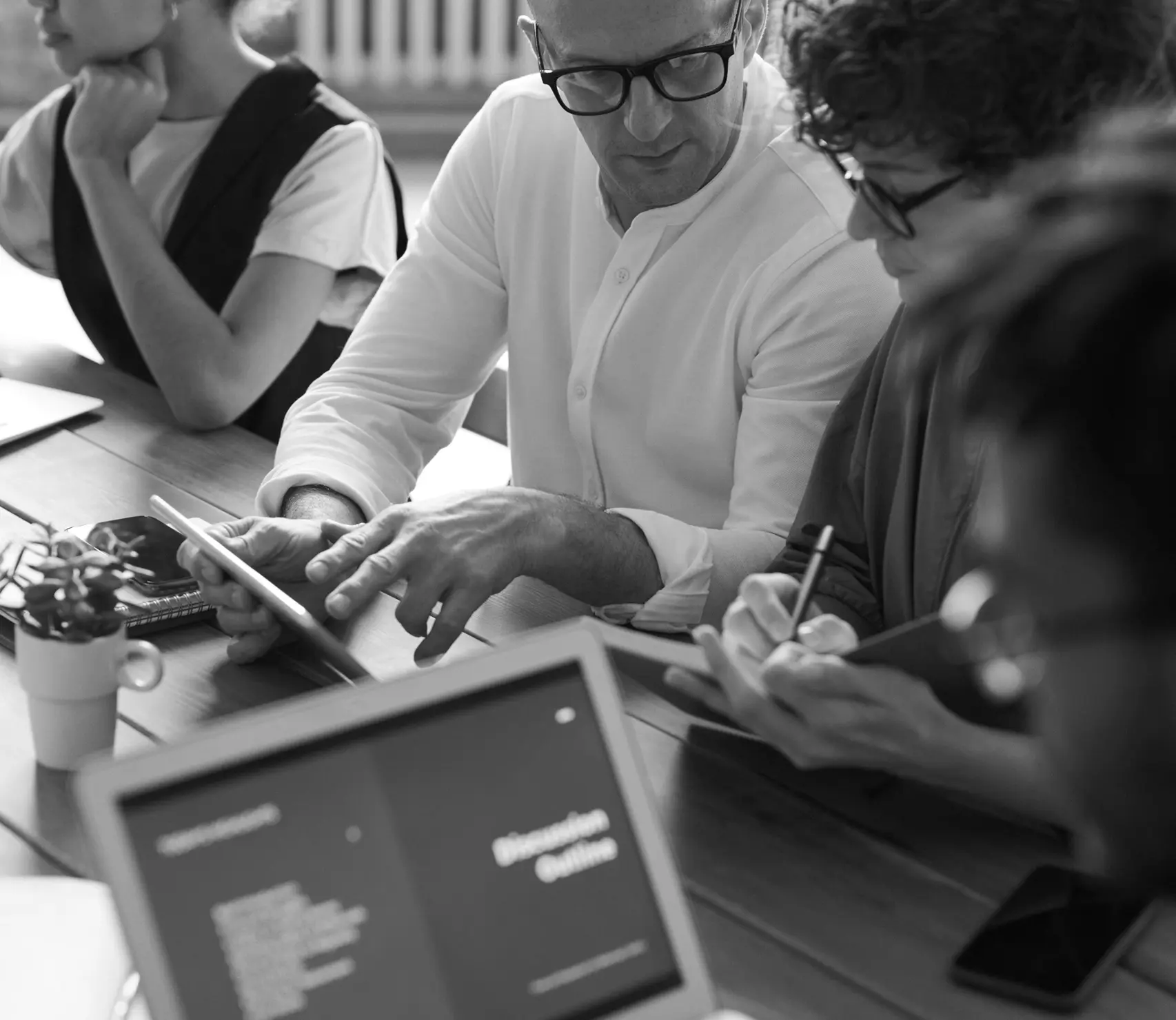 Group of four people collaborating around a table with laptop, tablet, and notebooks during a meeting.