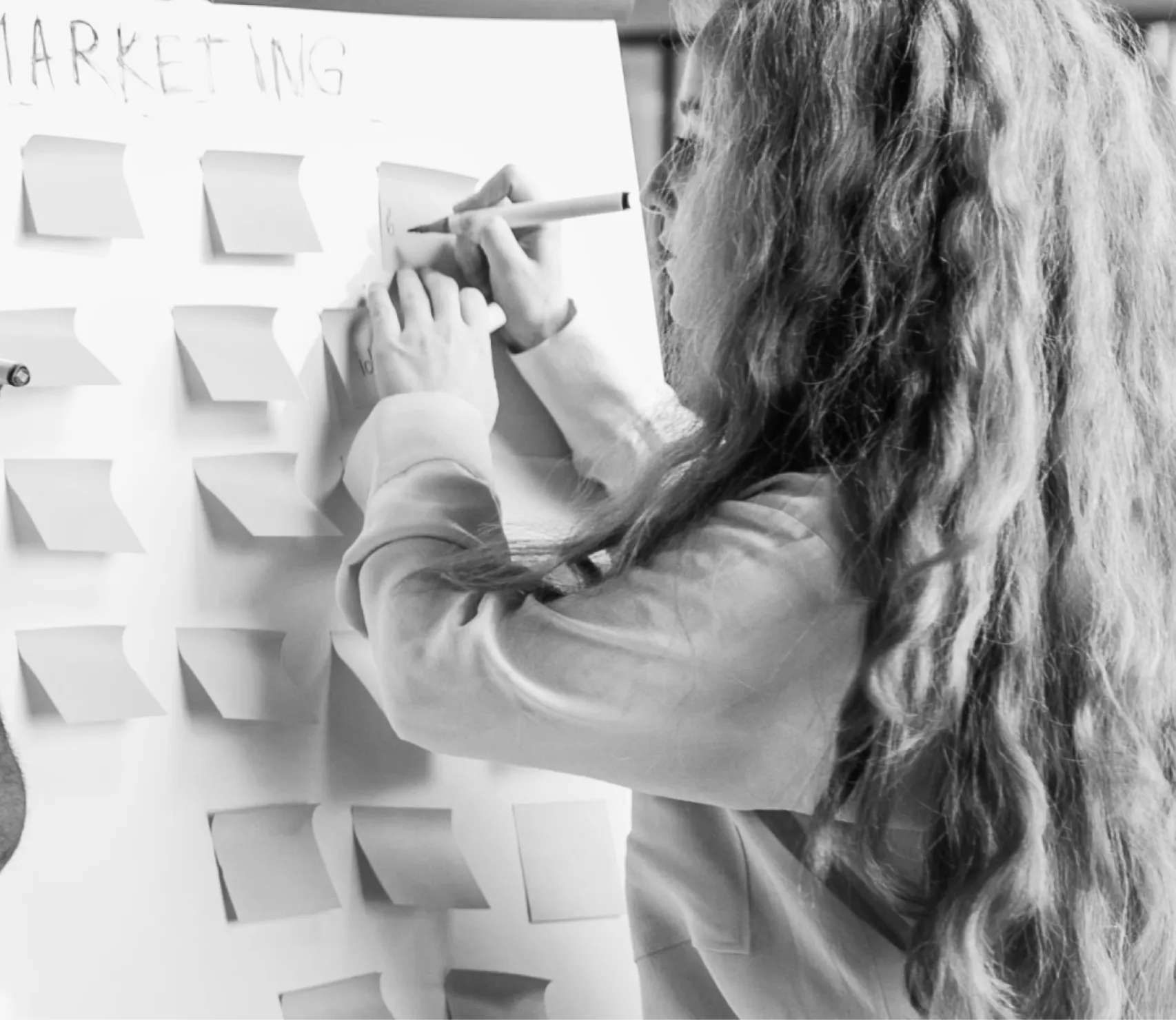 Person with long hair writing on sticky notes arranged on a board labeled 'MARKETING'.