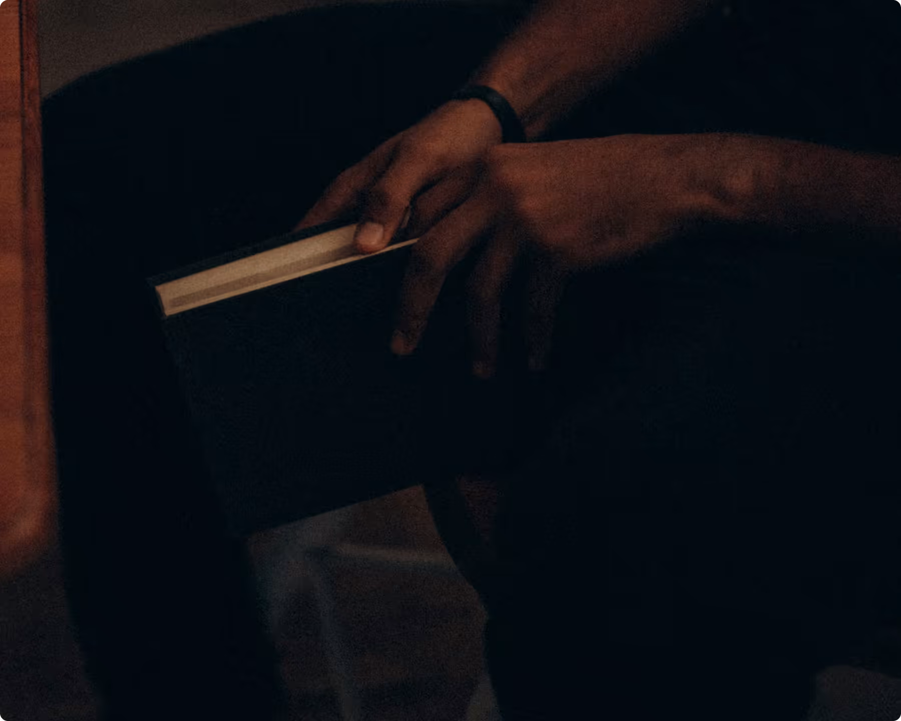 Dimly lit close-up of a person's hands holding a closed dark book, its pale page edges catching the light.