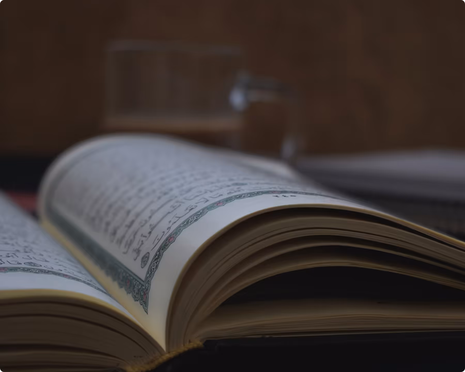 Close-up of an open book with Arabic script and a decorative green border on cream-colored pages, with a blurred glass cup in the background.
