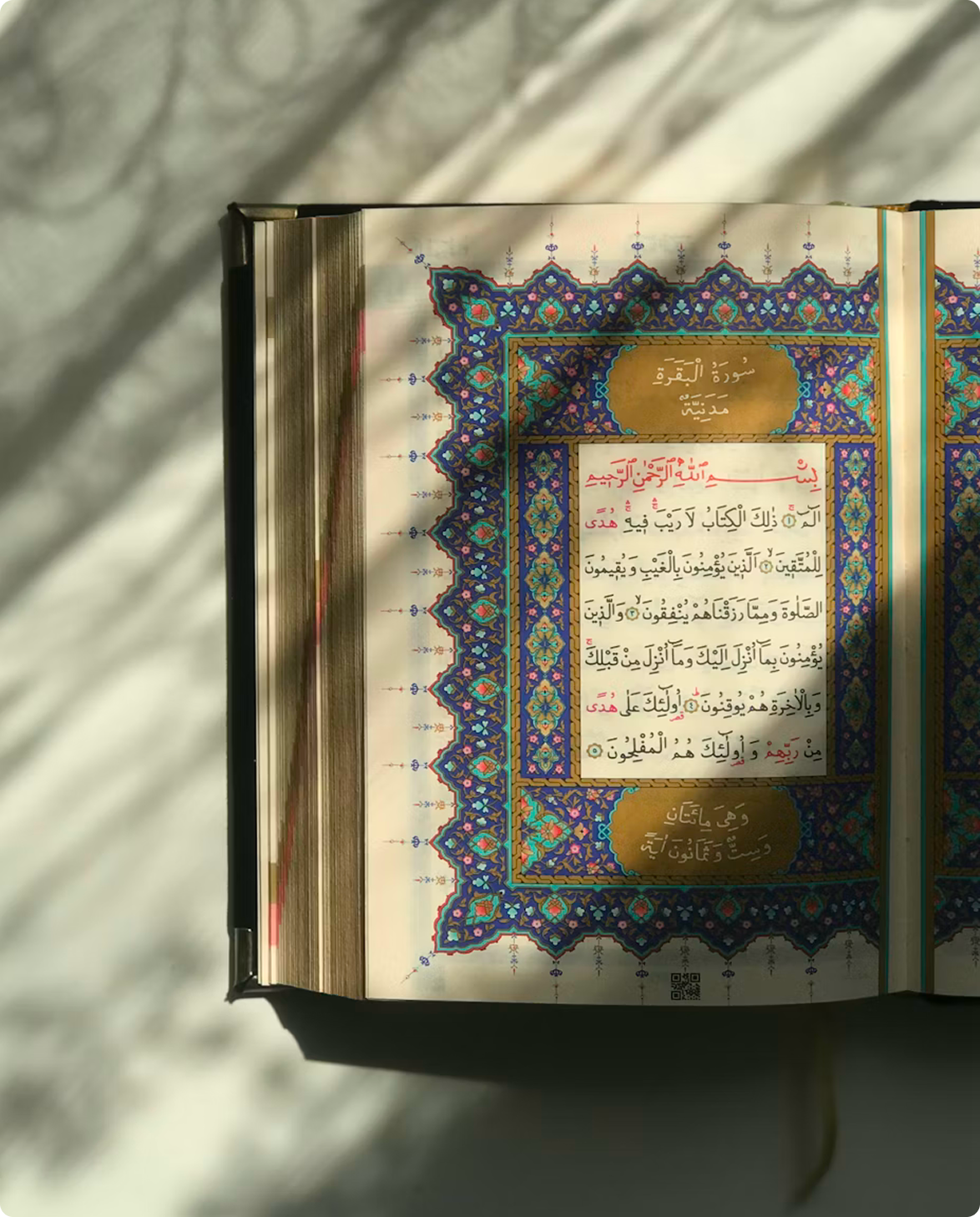 Open Quran showing an ornate, illuminated page with Arabic calligraphy and an intricate blue, purple and gold geometric border.