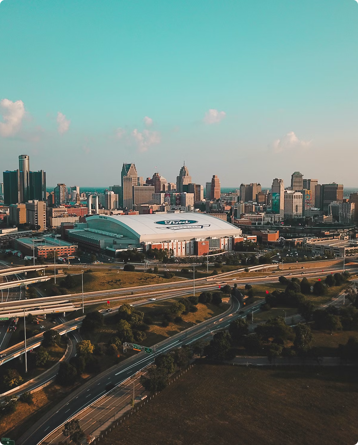 Aerial view of Detroit's downtown skyline and Ford Field stadium behind interlocking highways in warm evening light.