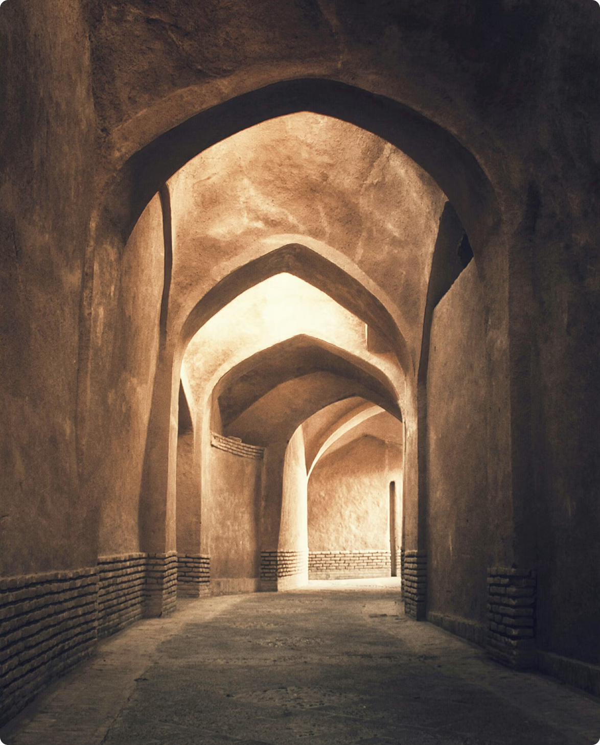 Dimly lit passage of pointed mud-brick arches leading toward a bright opening.