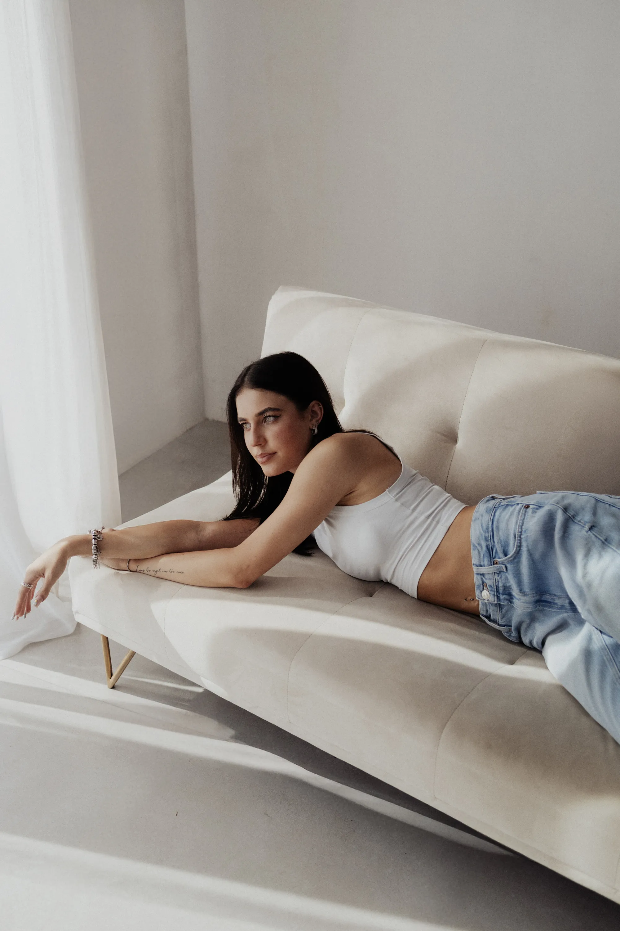 Modelka leżąca na jasnej kanapie w białym topie, naturalne oświetlenie w studio.