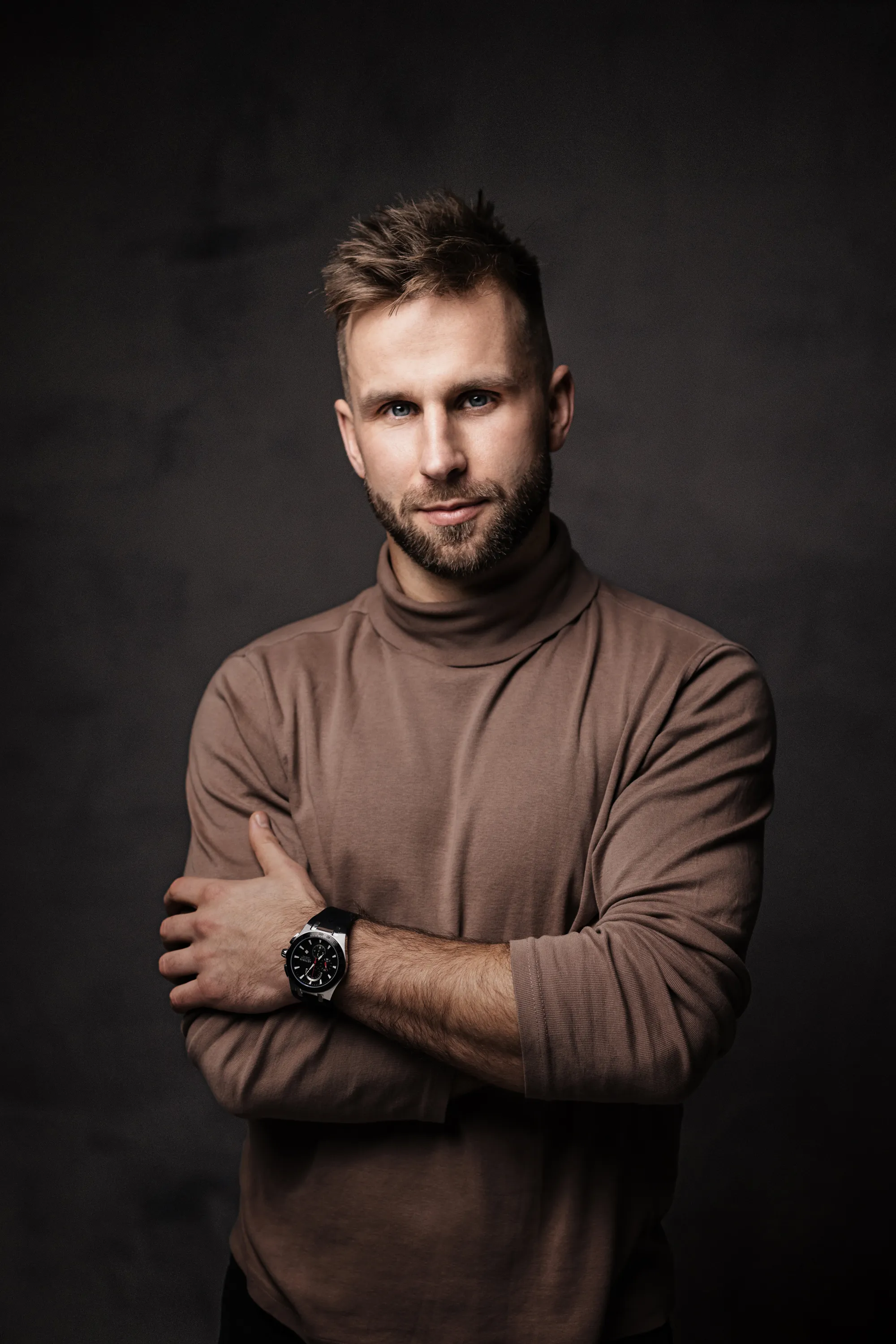 Studyjny portret męski – mężczyzna z założonymi rękami w brązowym golfie na ciemnym tle.
