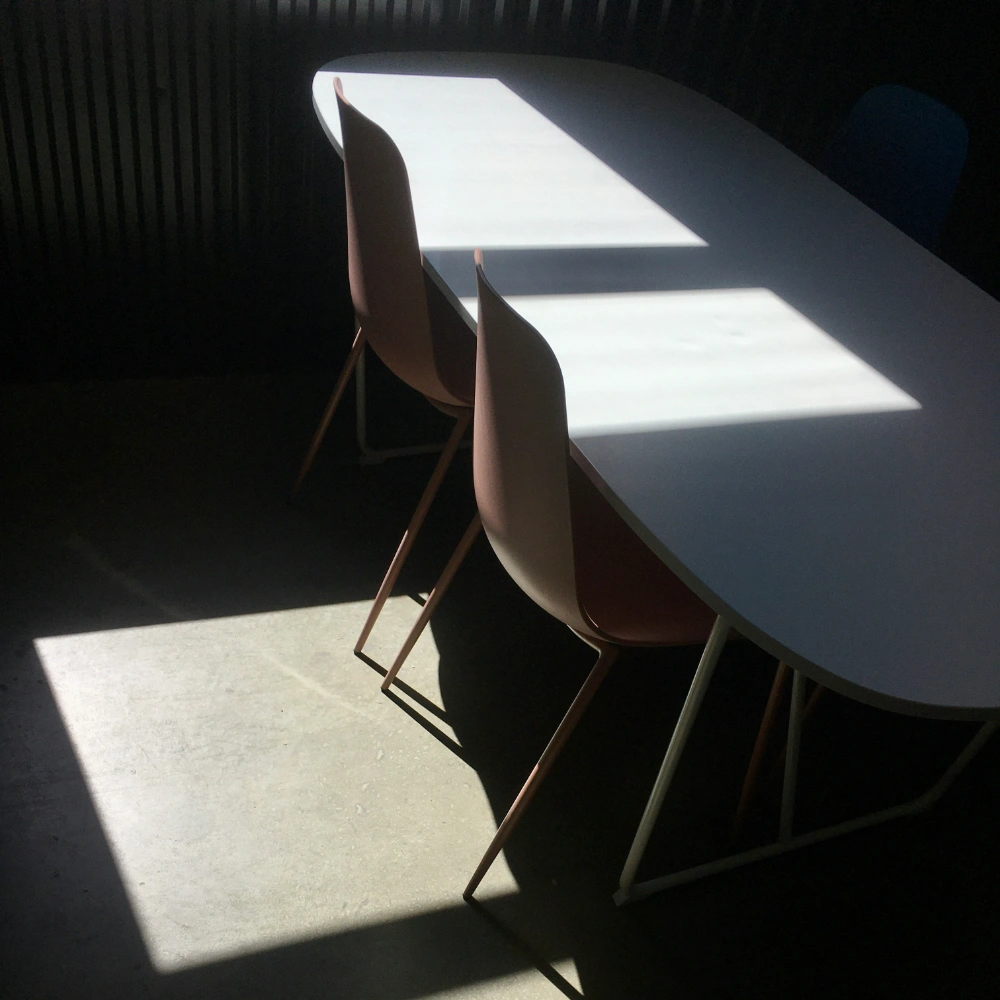 Minimalistický interiér s bielym stolom, stoličkami a výraznými tieňmi svetla.