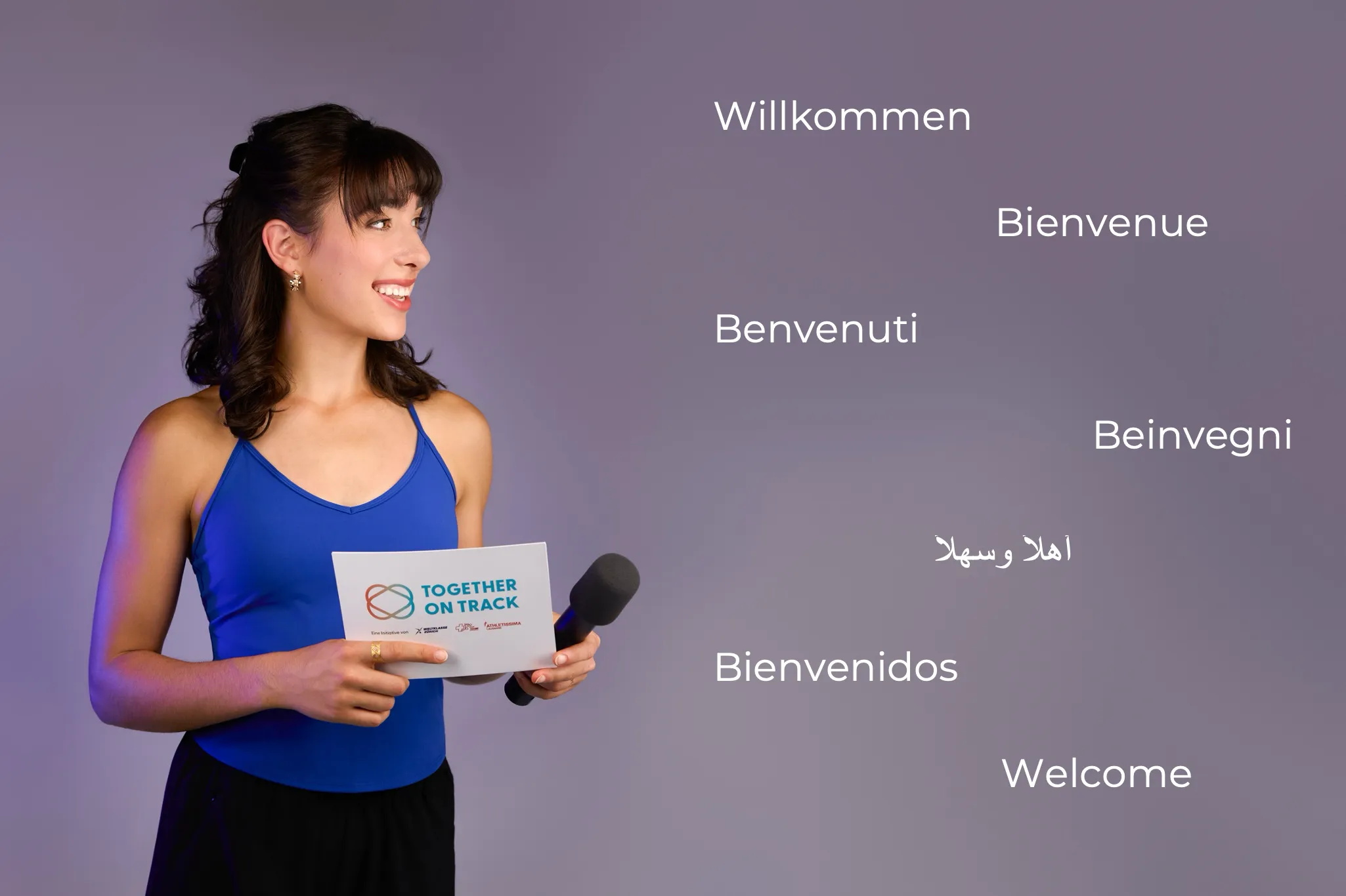 Ladina Ziani with a microphone and a moderation card, greeting in seven languages: Willkommen, Bienvenue, Benvenuti, Beinvegni, Ahlan wa sahlan, Bienvenidos, Welcome