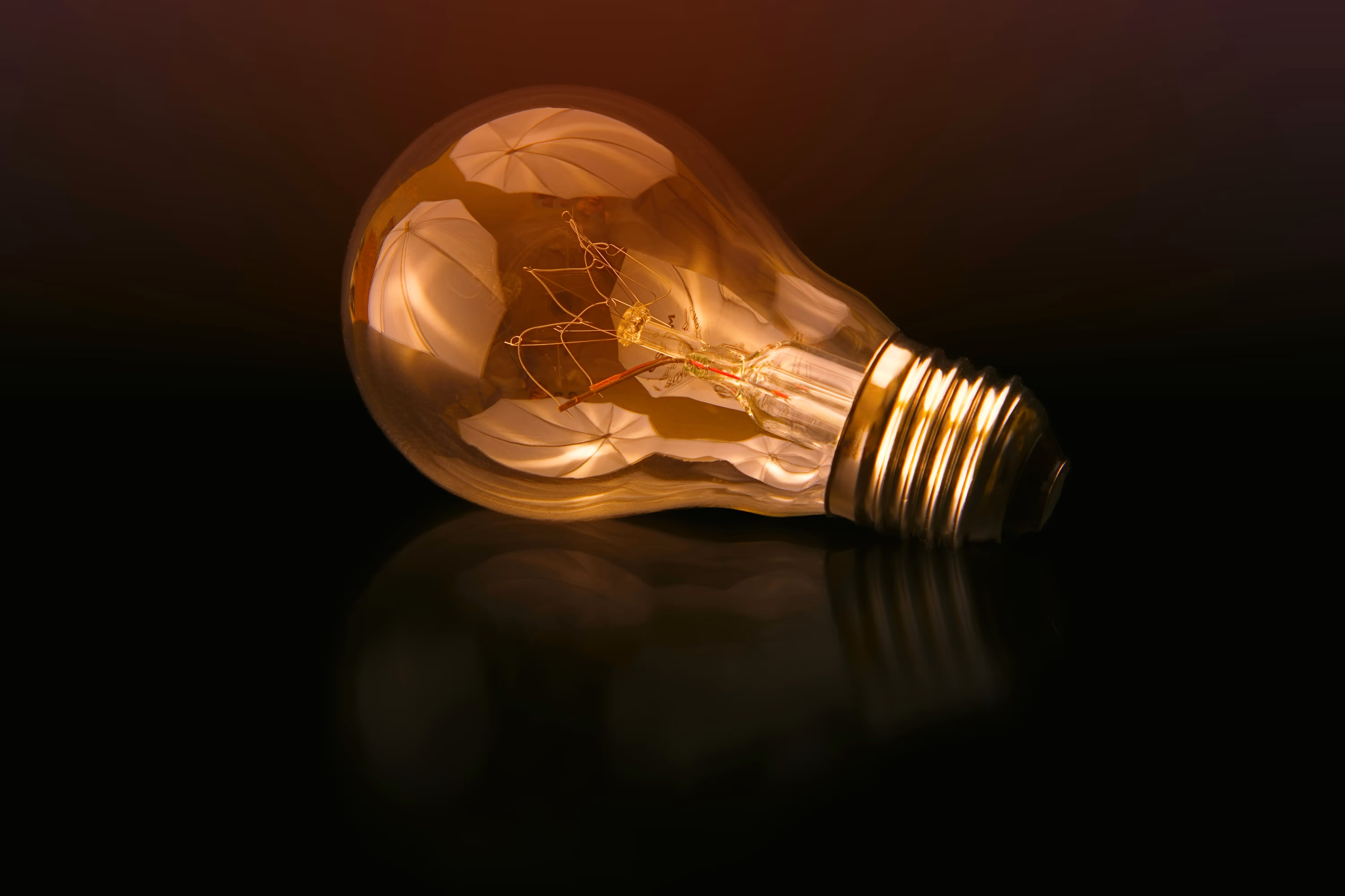 Close-up of a light bulb lying on a reflective black surface with warm light reflections inside.