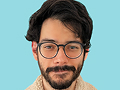 Man with dark hair, beard, and glasses wearing a light brown shirt against a blue background.