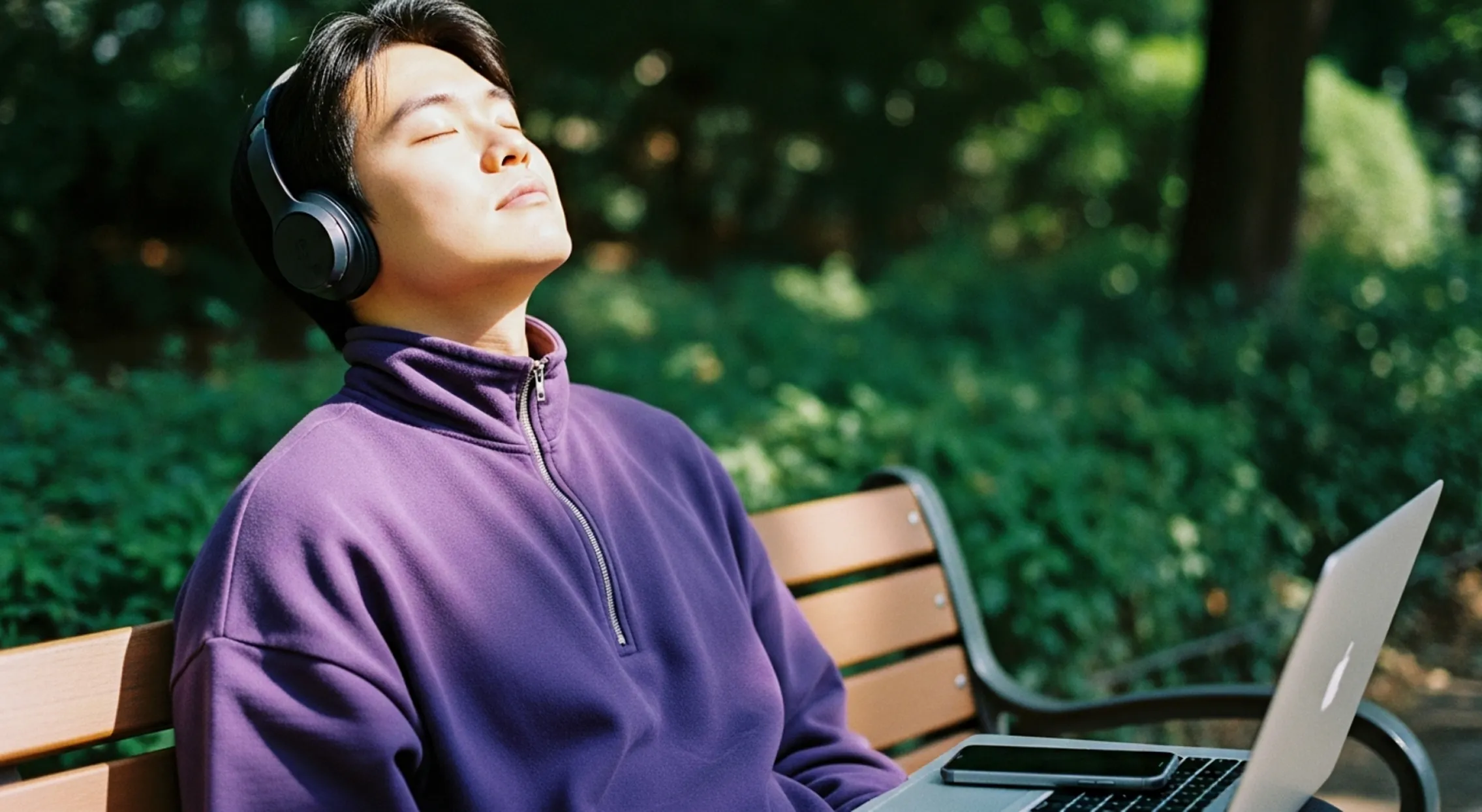 Person wearing headphones and a purple jacket, sitting on a bench with a laptop and phone nearby, eyes closed.
