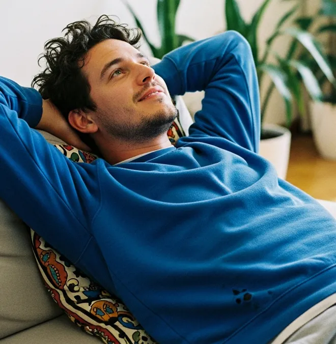 Relaxed young man reclining on couch with hands behind head, looking upward, with plants in the background.