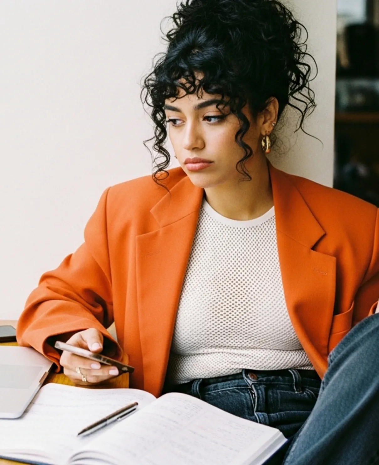 Woman in orange blazer holding a phone, sitting beside an open notebook and laptop, looking to the side.