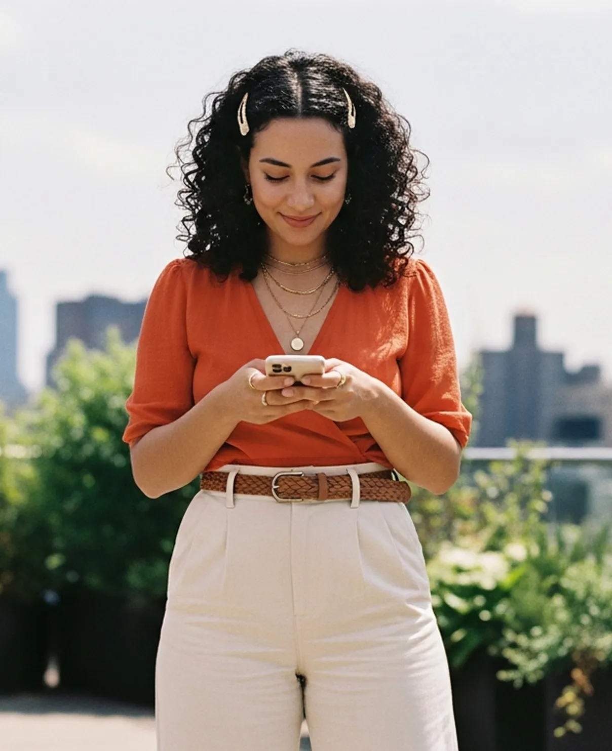 Woman with curly hair wearing an orange top and white pants, looking down at her smartphone outdoors.