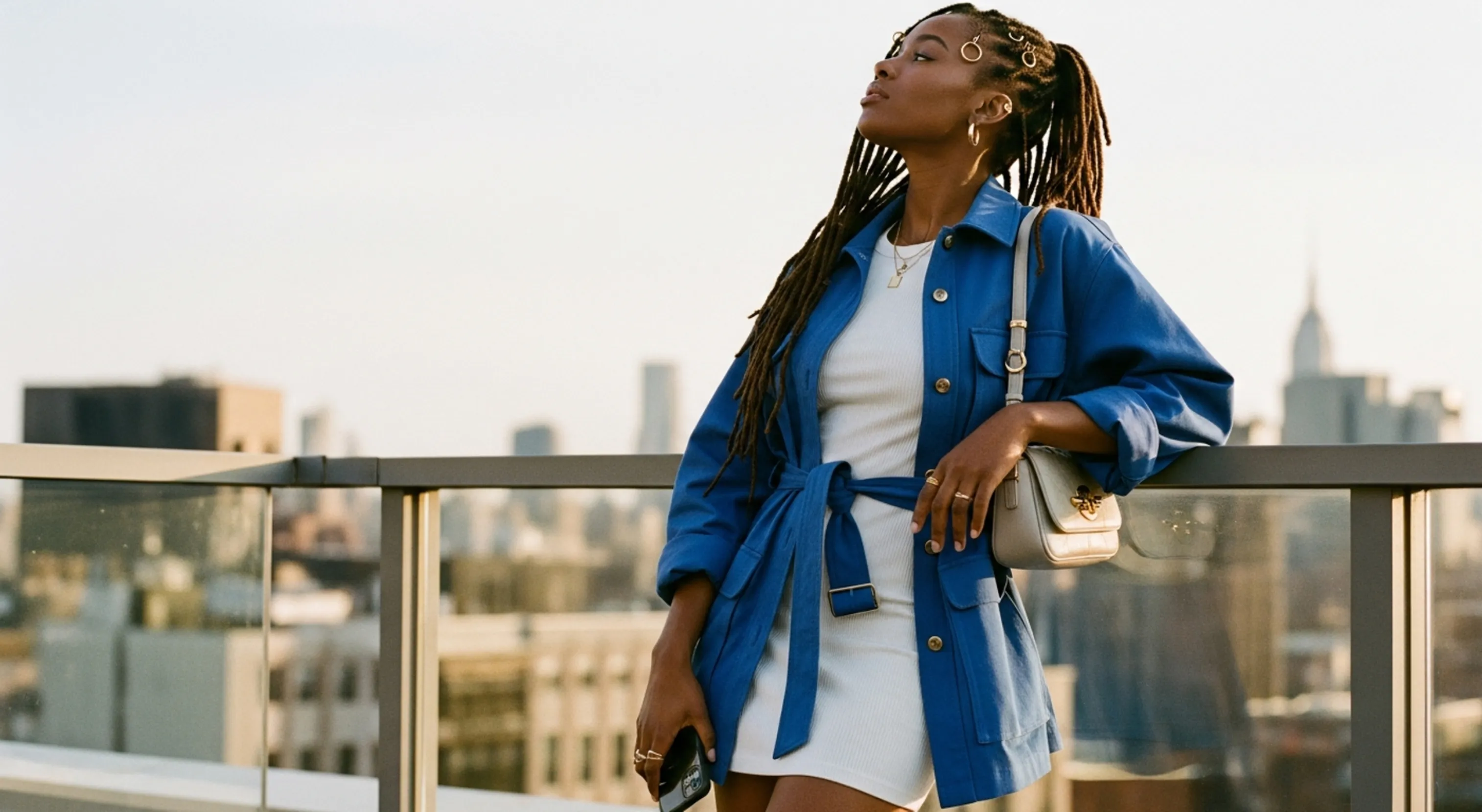 Woman with dreadlocks in blue jacket and white dress standing on rooftop with city skyline behind her.