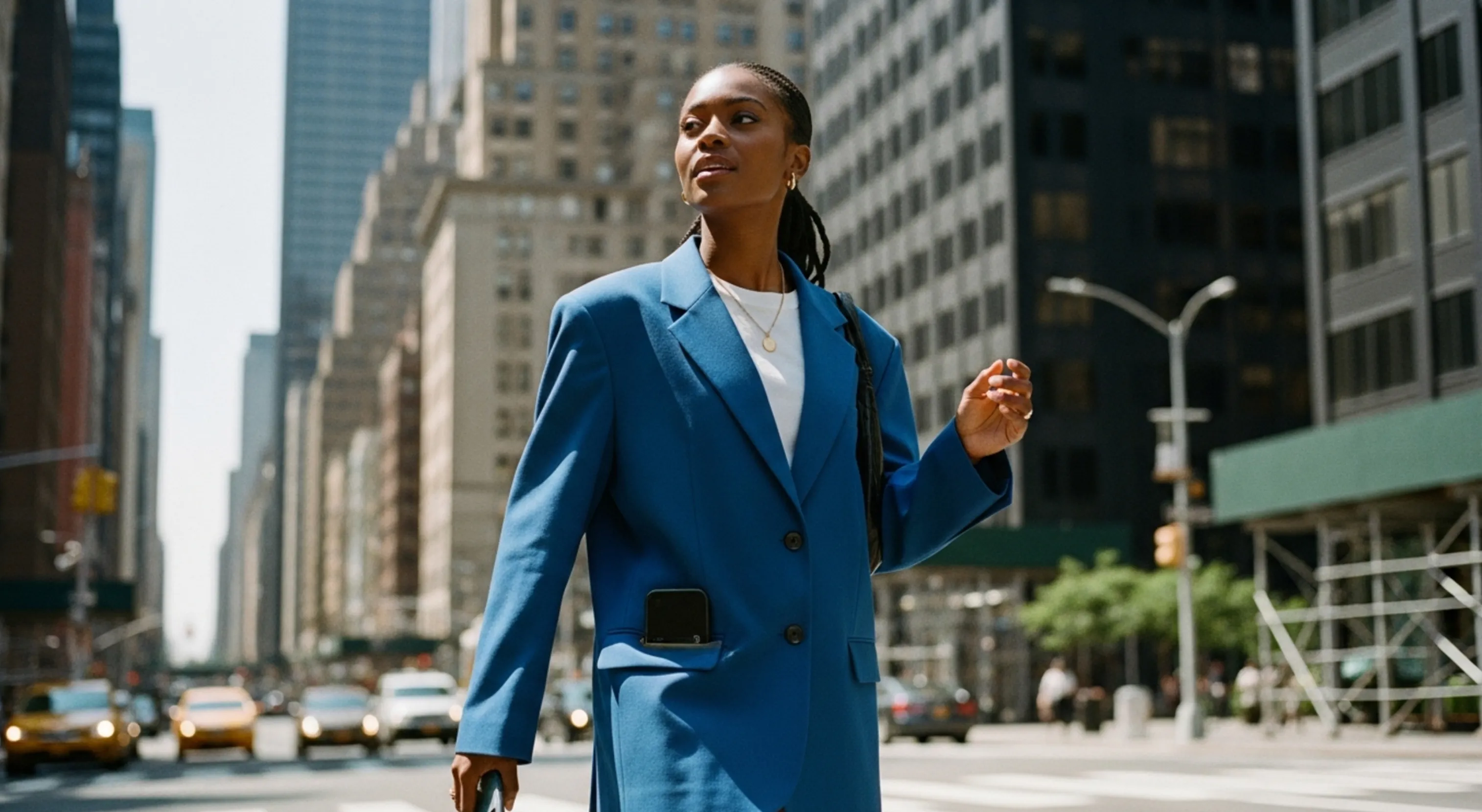 Woman in a blue blazer walking confidently on a city street with buildings and cars behind her.