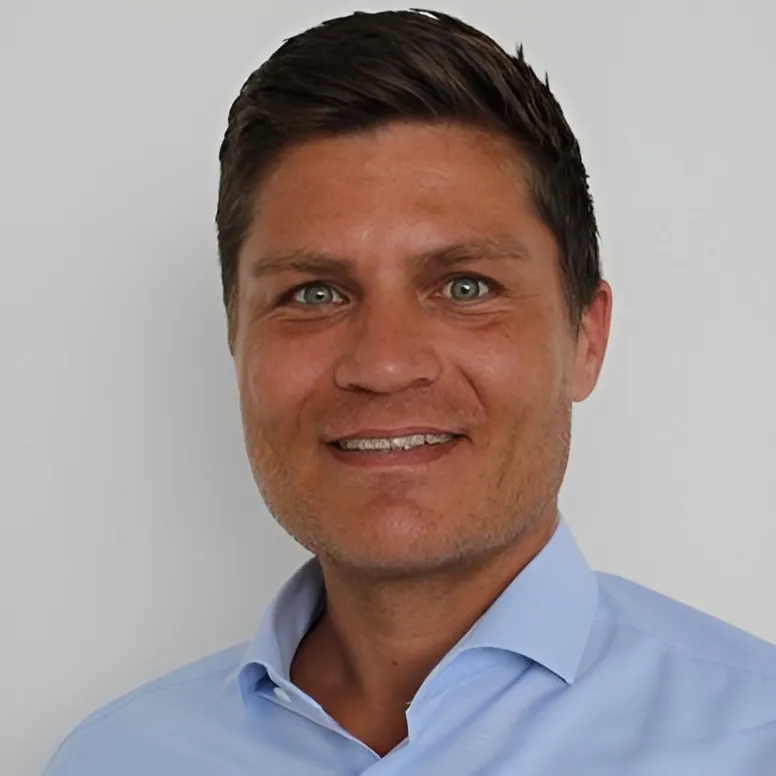 Portrait of a man with short dark hair and light blue eyes, wearing a light blue collared shirt, smiling gently against a plain light gray background.