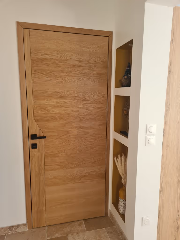 Porte intérieure en bois clair avec poignée noire à gauche dans un mur blanc avec étagères encastrées décorées.