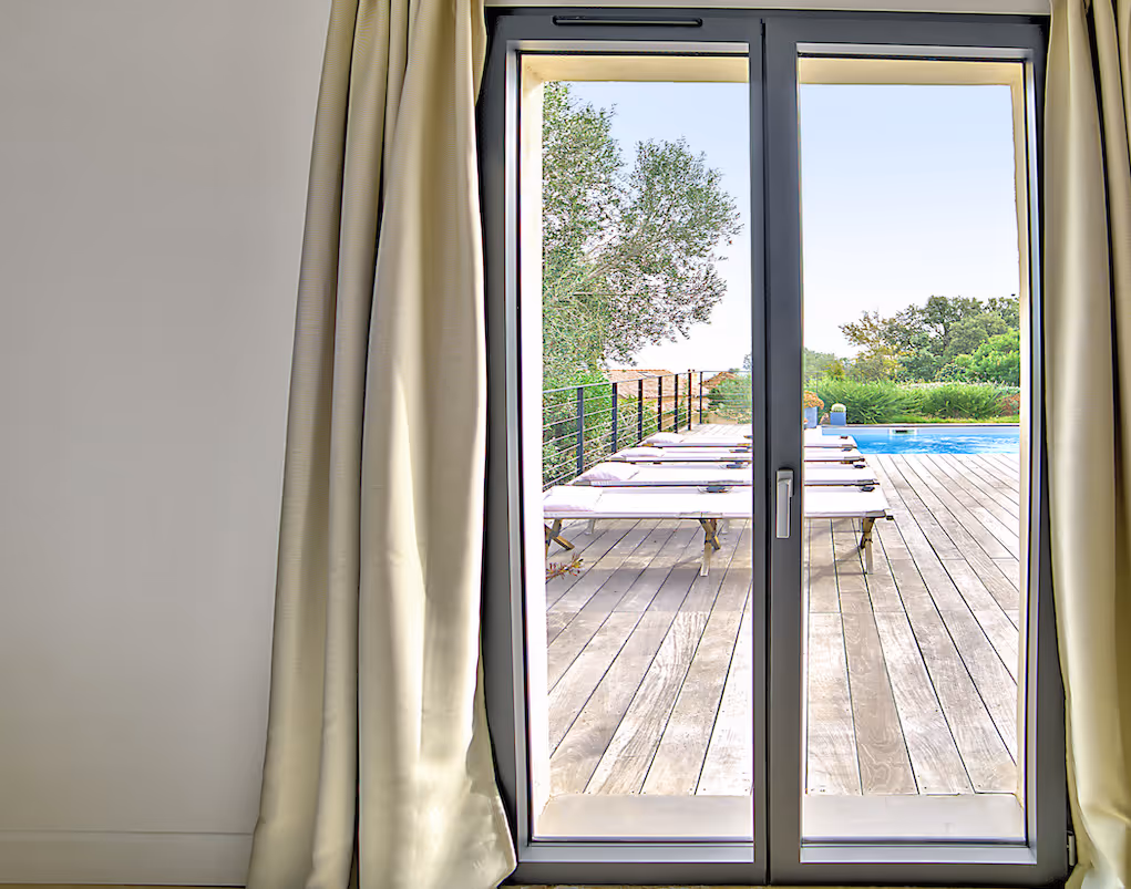 Vue à travers une porte-fenêtre vers une terrasse en bois avec des transats alignés et une piscine, entourée d'arbres verts.