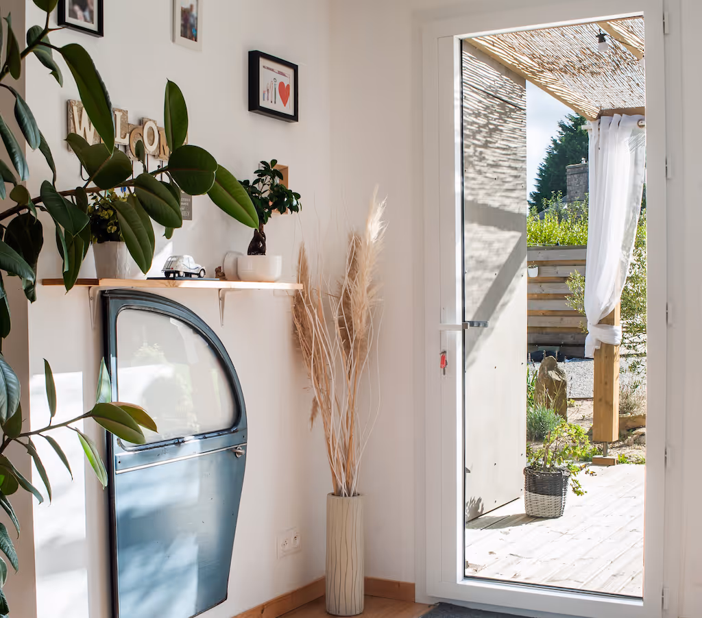 Entrée lumineuse avec porte en verre donnant sur une terrasse, décorée avec une vieille porte de voiture bleue fixée au mur, des plantes et un vase avec des herbes sèches.