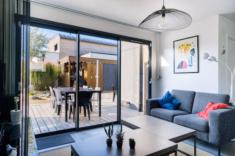 Salon moderne avec canapé gris, coussins colorés et table basse, vue sur terrasse en bois avec table et chaises par portes vitrées coulissantes.