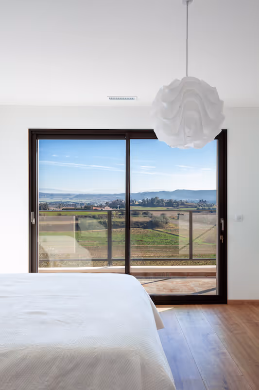 Chambre minimaliste avec lit blanc face à une grande baie vitrée offrant une vue sur un paysage champêtre et des collines au loin.