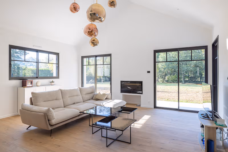 Salon lumineux avec canapé beige, tables basses en verre, cheminée moderne et grandes fenêtres donnant sur un jardin verdoyant.