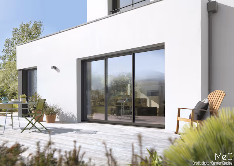 Maison moderne blanche avec grandes fenêtres coulissantes donnant sur une terrasse en bois équipée de meubles de jardin et de plantes.