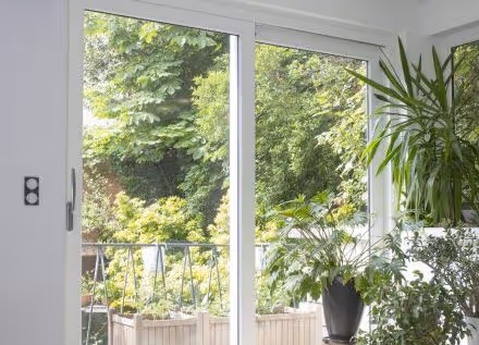 Porte-fenêtre coulissante en bois avec vue sur un balcon entouré de plantes vertes et d'arbres feuillus.