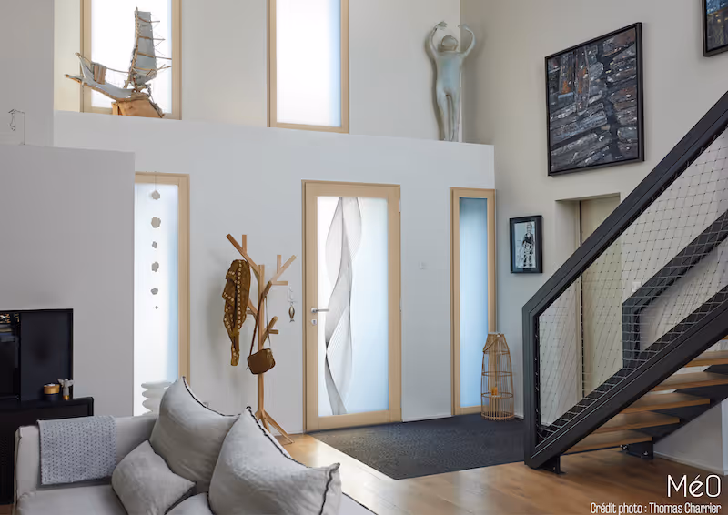 Intérieur moderne avec canapé clair, porte en verre artistique, patère en bois, escalier avec rampe métallique et œuvres d'art accrochées.