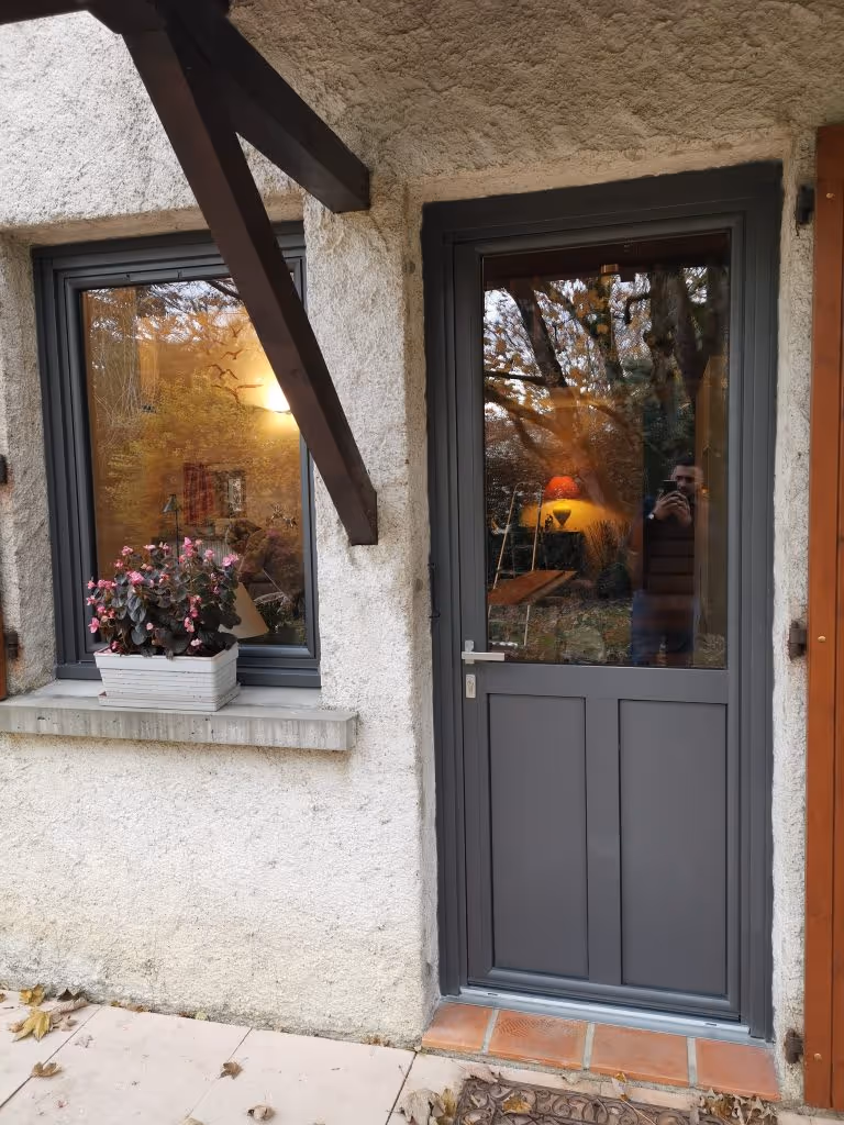Porte grise avec une grande vitre et une fenêtre adjacente avec jardinière fleurie sur un mur en crépi clair.