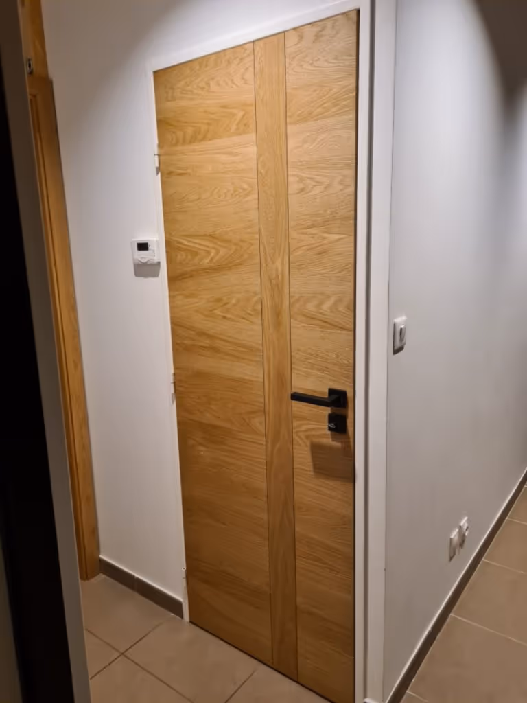 Porte intérieure en bois clair avec poignée noire dans un couloir aux murs blancs et sol carrelé beige.